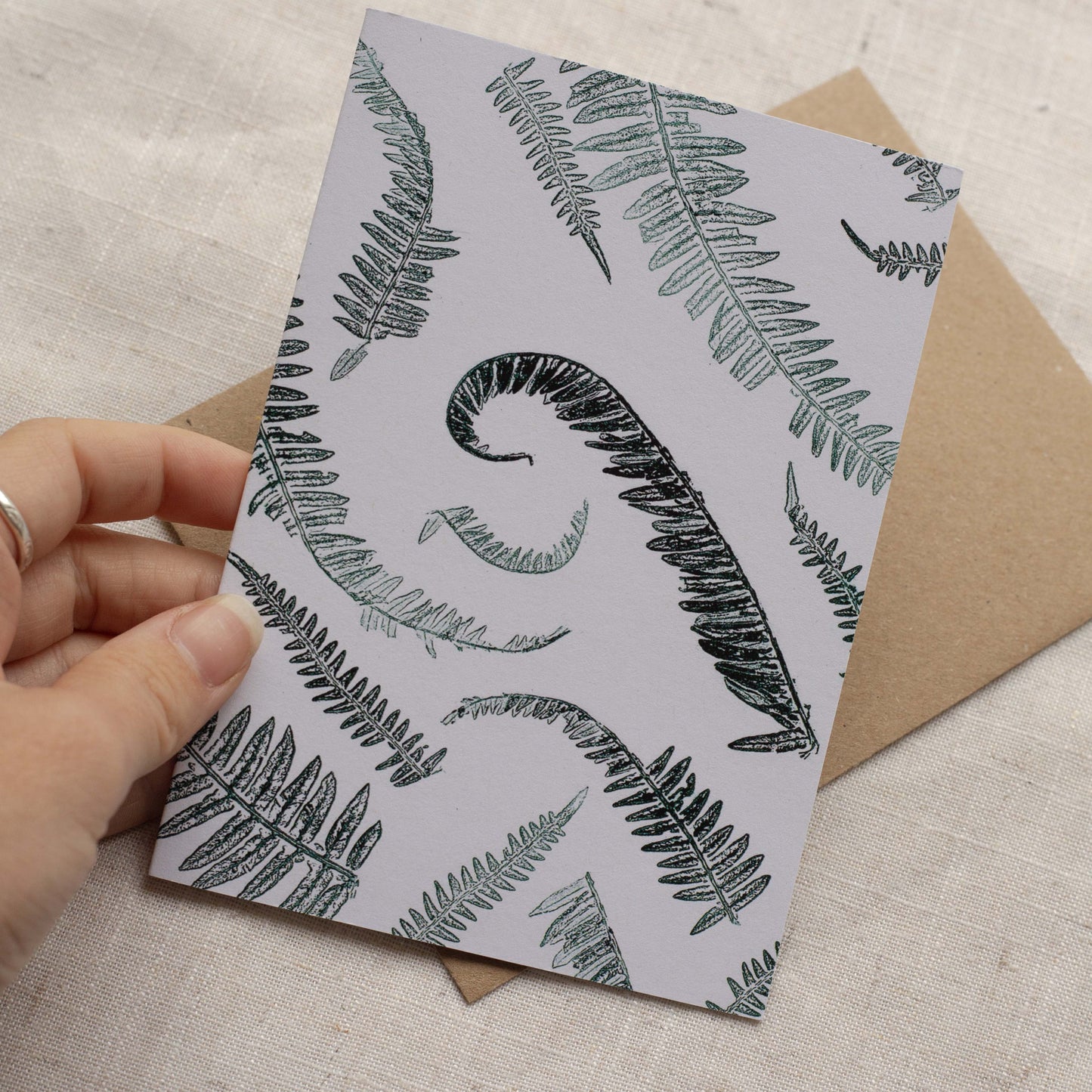 Card with fern pattern held by a hand, with a brown envelope on a textured surface