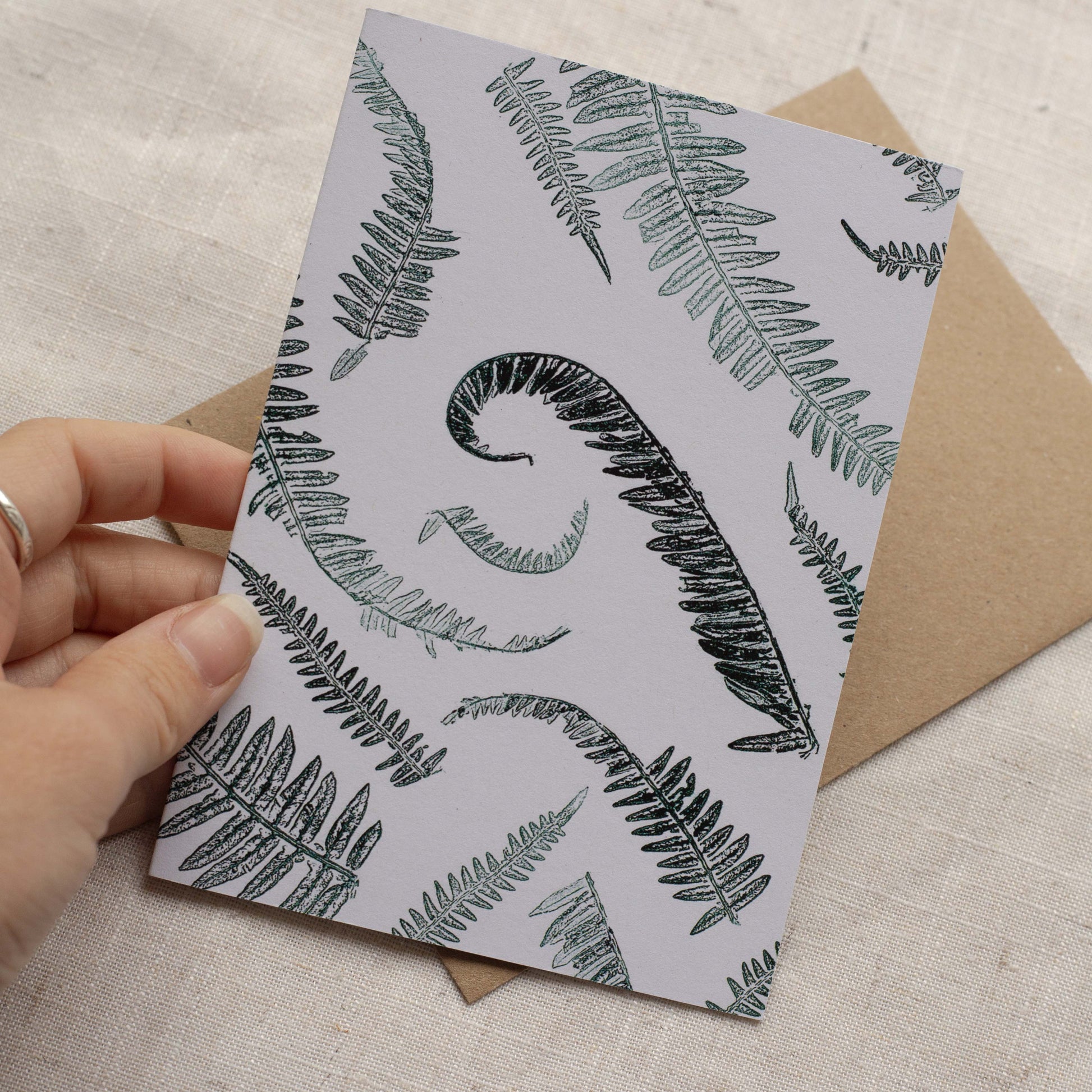 Card with fern pattern held by a hand, with a brown envelope on a textured surface