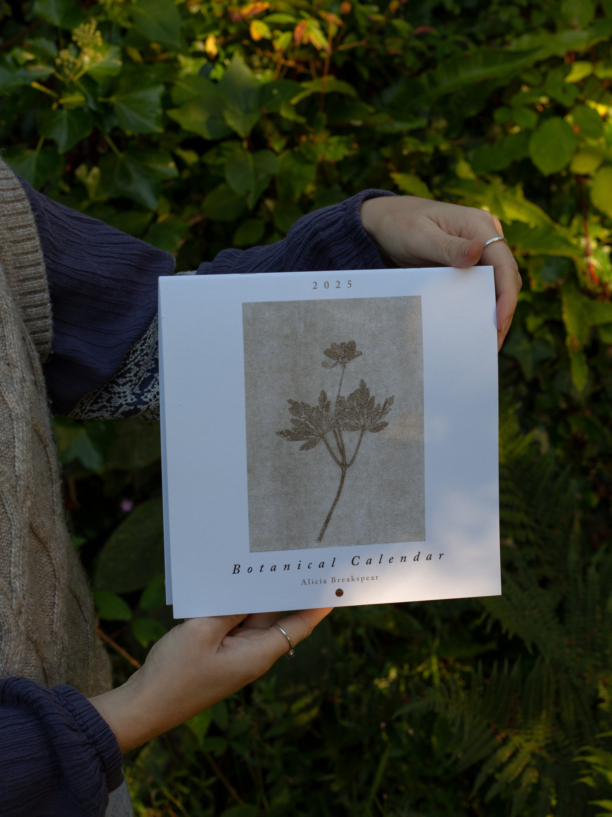 Outside with the 2025 botanical calendar being held by Alicia, the artist.