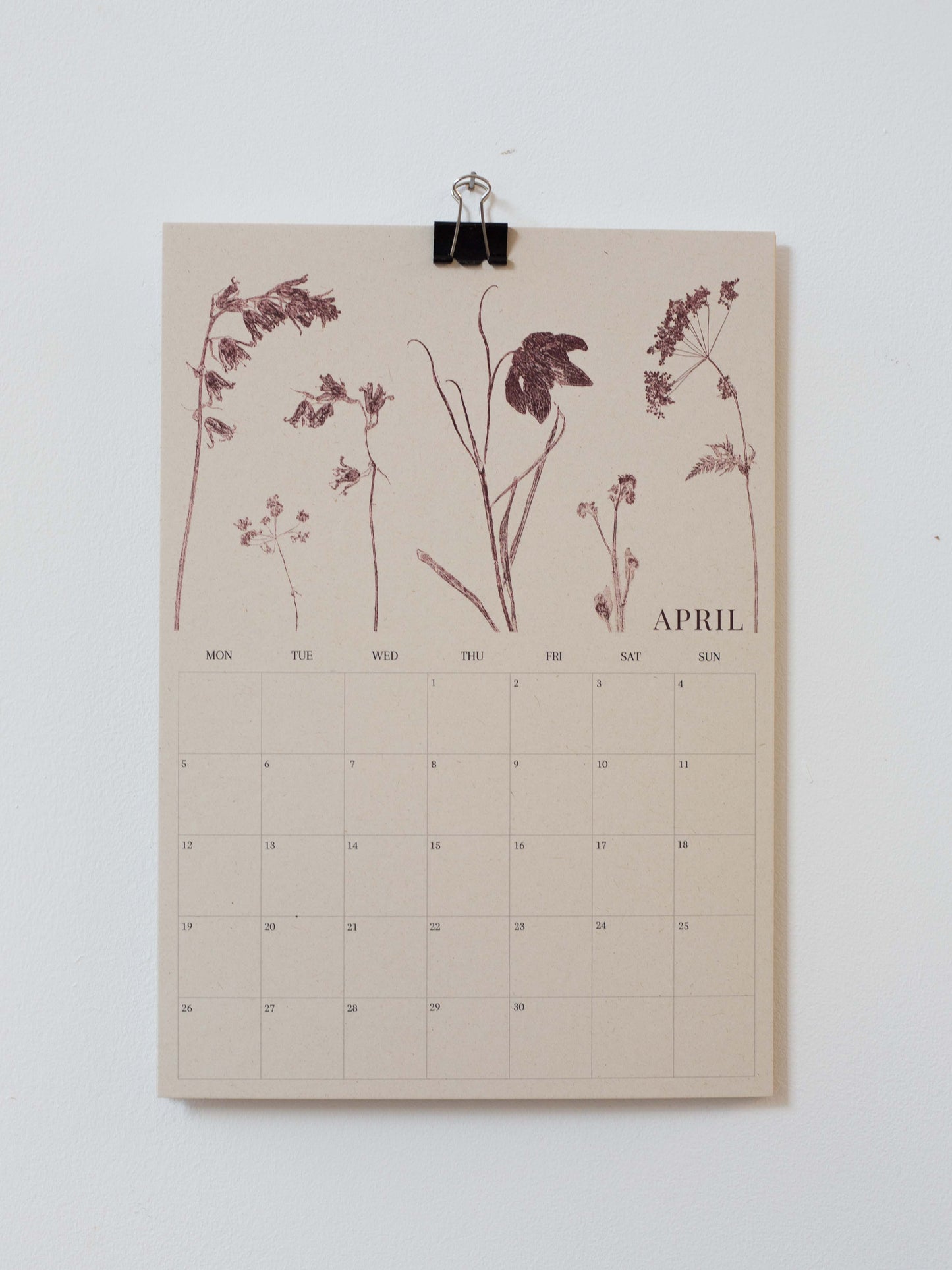 A beige April wall calendar page with botanical illustrations in dark brown, clipped at the top with a black binder clip against a plain white wall.