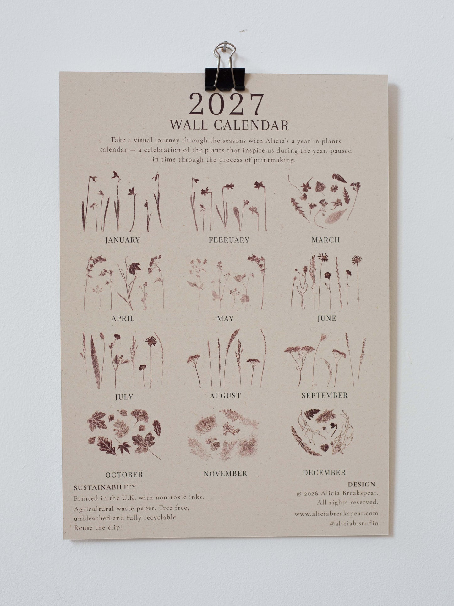 A 2027 wall calendar printed on a beige paper sheet, clipped at the top with a metal clip against a light gray wall. The calendar features botanical illustrations and text for each month, with sustainability and design credits at the bottom.