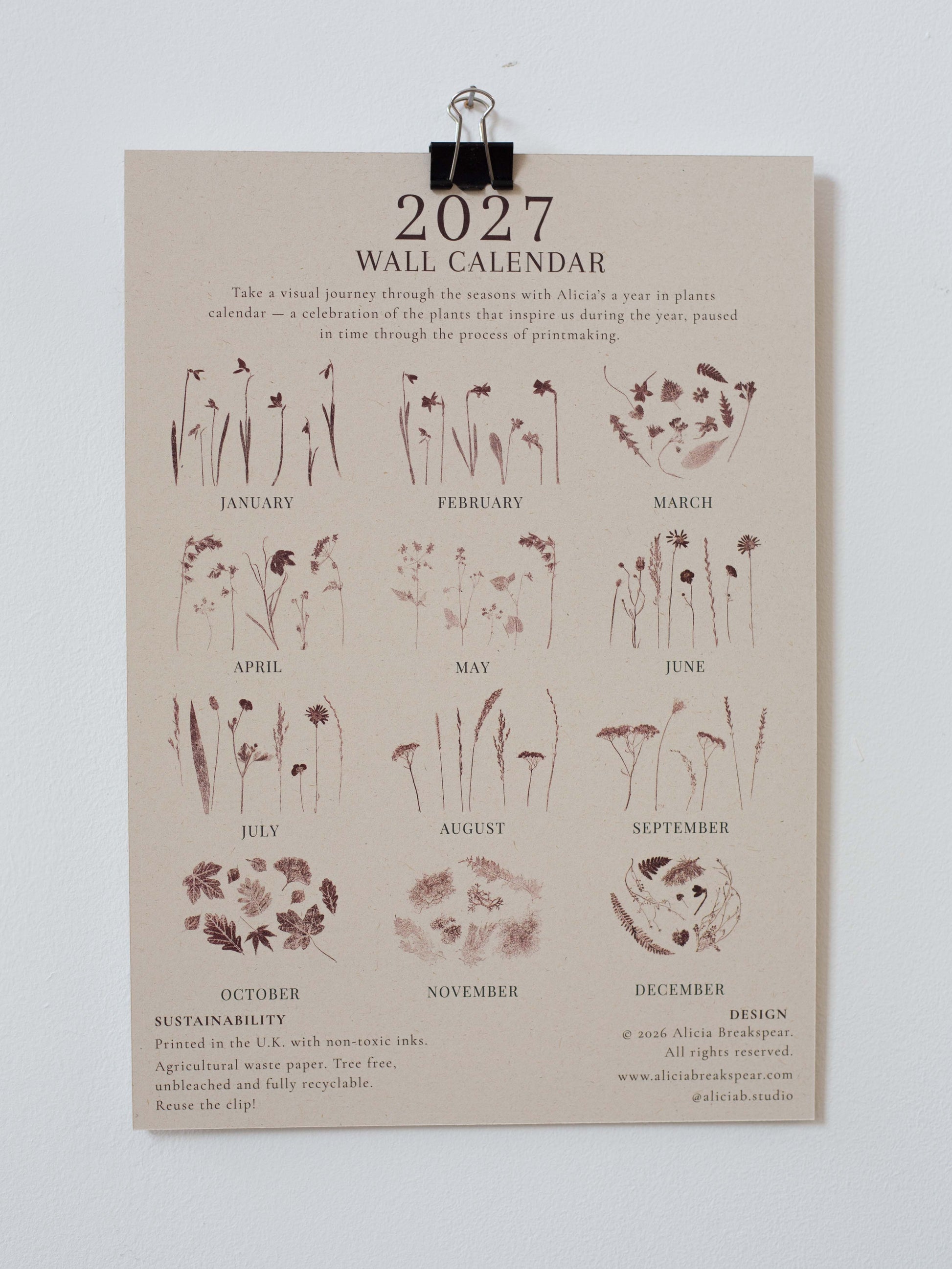 A 2027 wall calendar printed on a beige paper sheet, clipped at the top with a metal clip against a light gray wall. The calendar features botanical illustrations and text for each month, with sustainability and design credits at the bottom.