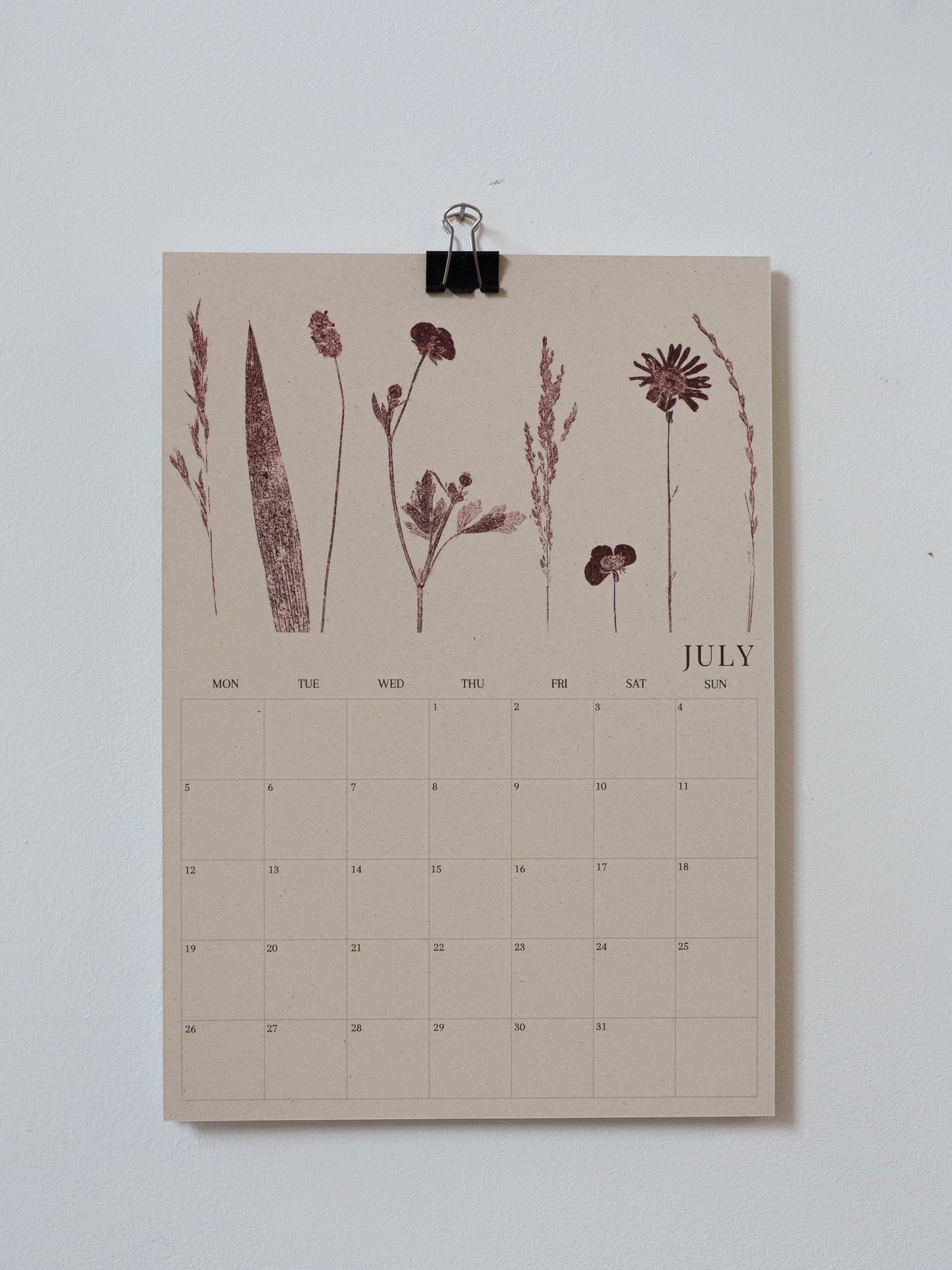 A minimalist July calendar page with botanical illustrations in muted burgundy on a warm beige background, clipped at the top with a black binder clip against a light gray wall.