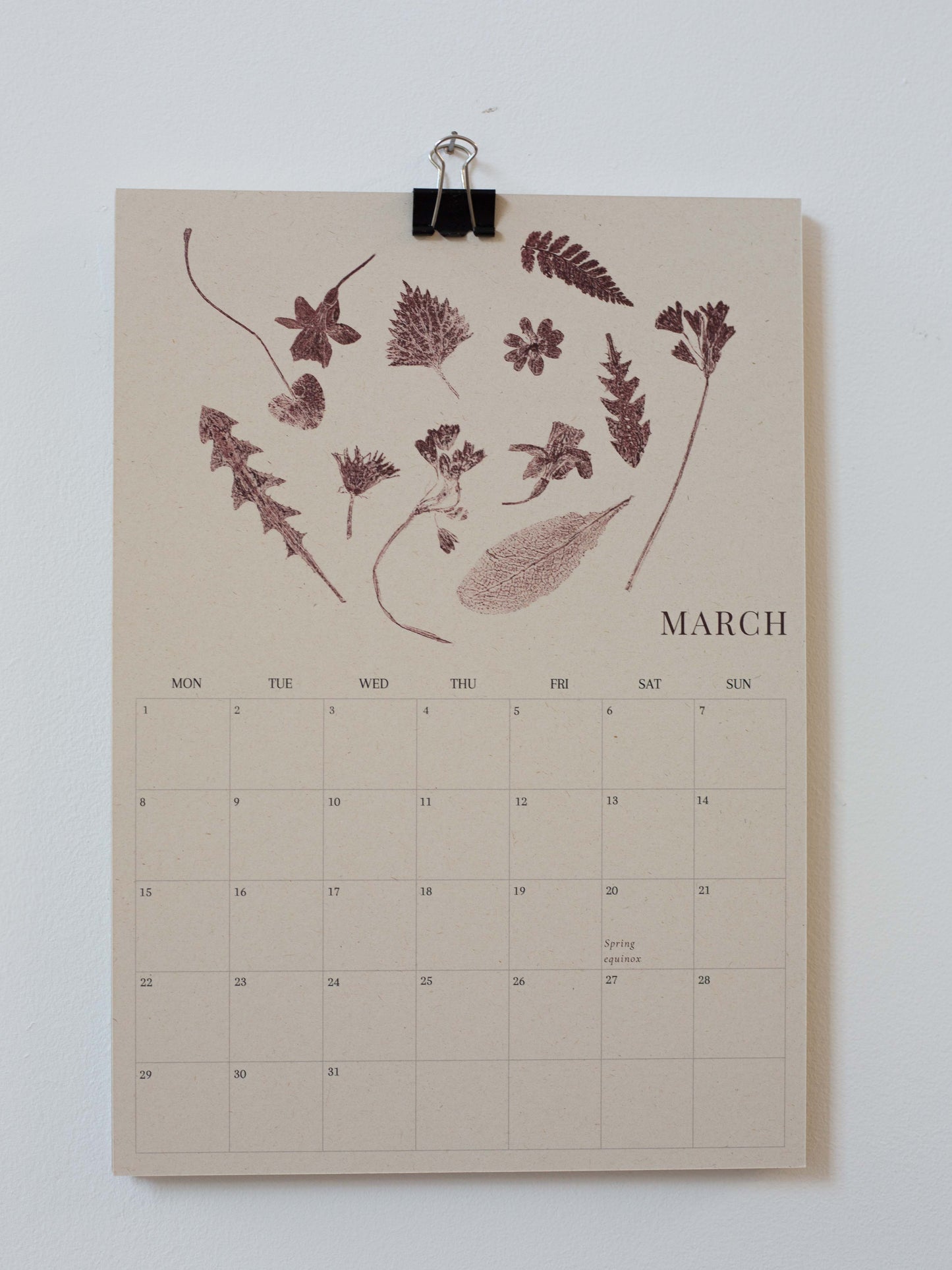 A beige monthly calendar page clipped at the top with a black binder clip, featuring botanical leaf illustrations across the top and the month name March with a numbered calendar grid below, shown against a plain light background.
