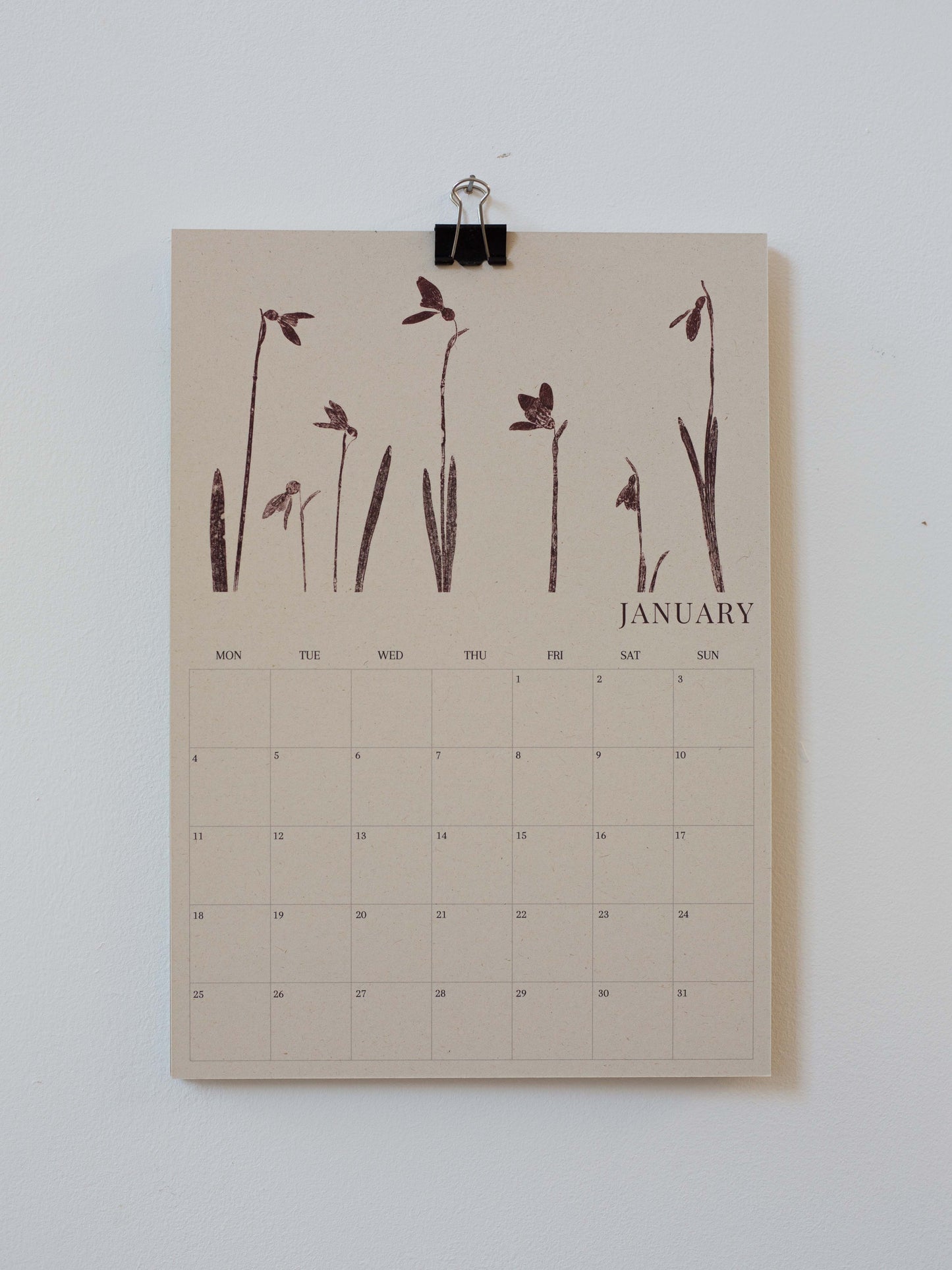 A minimalist January 2027 calendar page clipped at the top with a black binder clip against a plain white wall. The page has a warm beige background with dark brown botanical illustrations of snowdrops across the top and a grid calendar below labeled January.