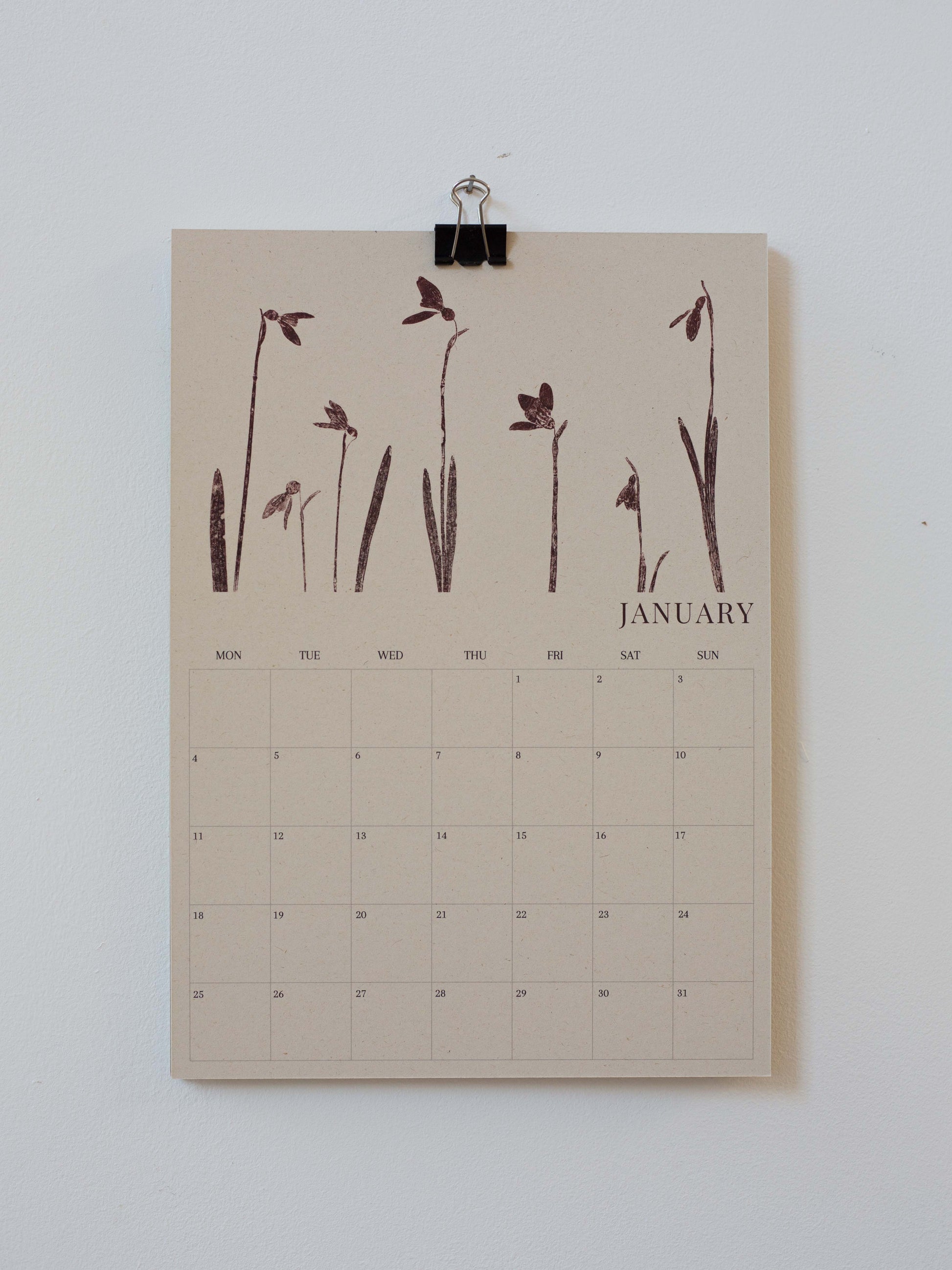 A minimalist January 2027 calendar page clipped at the top with a black binder clip against a plain white wall. The page has a warm beige background with dark brown botanical illustrations of snowdrops across the top and a grid calendar below labeled January.
