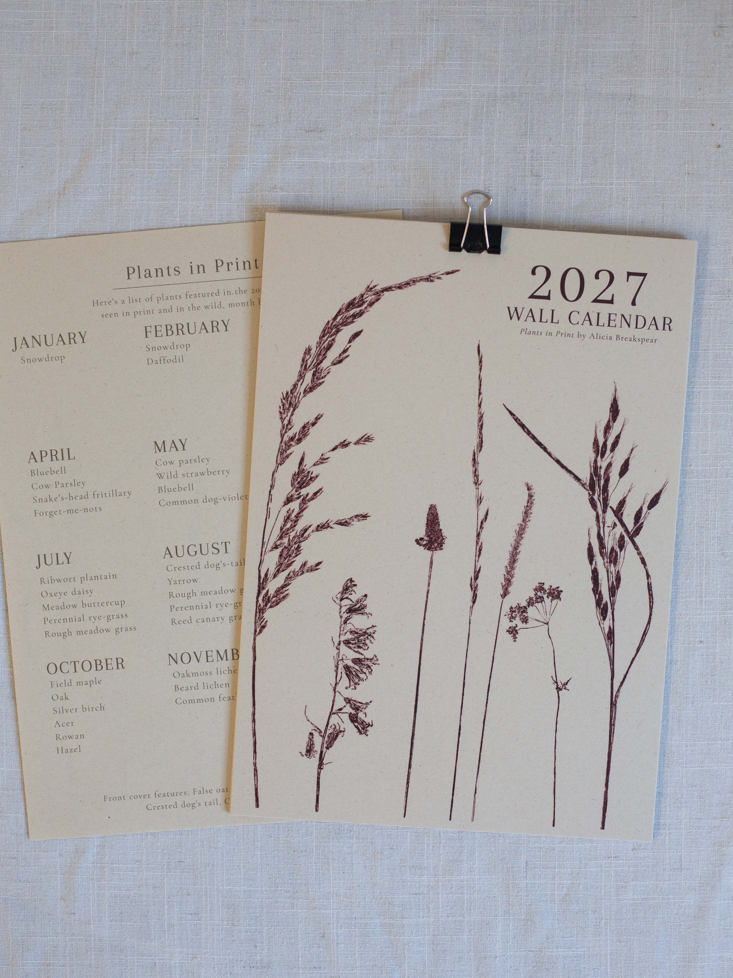 A 2027 wall calendar with botanical grass illustrations, displayed on a textured white linen background with a binder clip. A second calendar page showing plant lists is partially visible underneath.