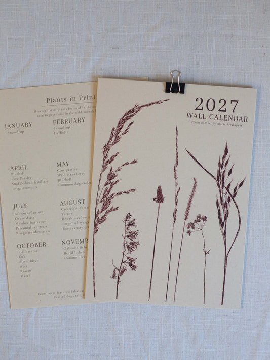 A 2027 wall calendar with botanical grass illustrations, displayed on a textured white linen background with a binder clip. A second calendar page showing plant lists is partially visible underneath.