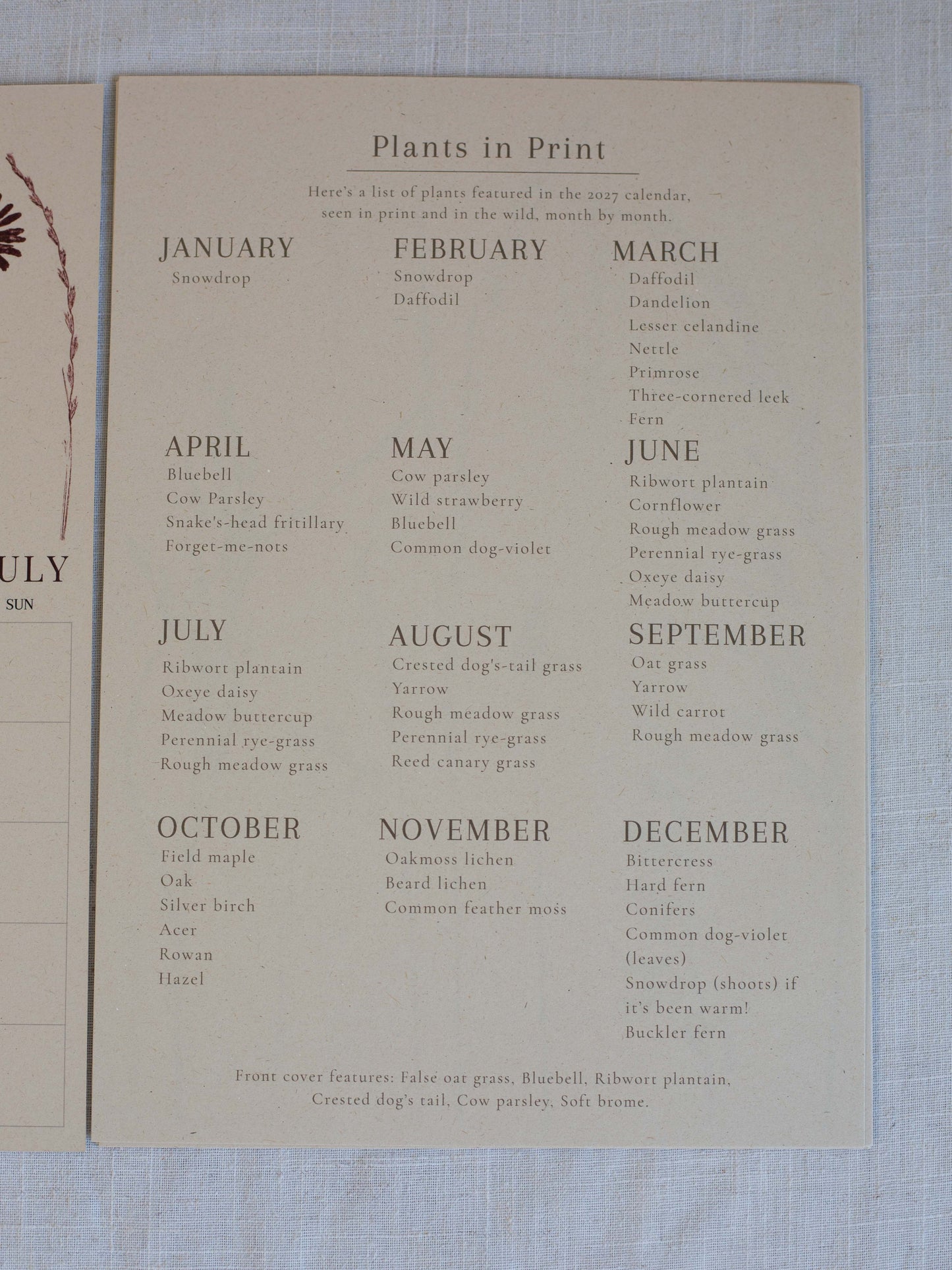 A vintage paper calendar page titled 'Plants in Print' laid on a light blue textured fabric background. The page lists months from January to December with plant names, and part of another paper sheet is visible on the left edge.