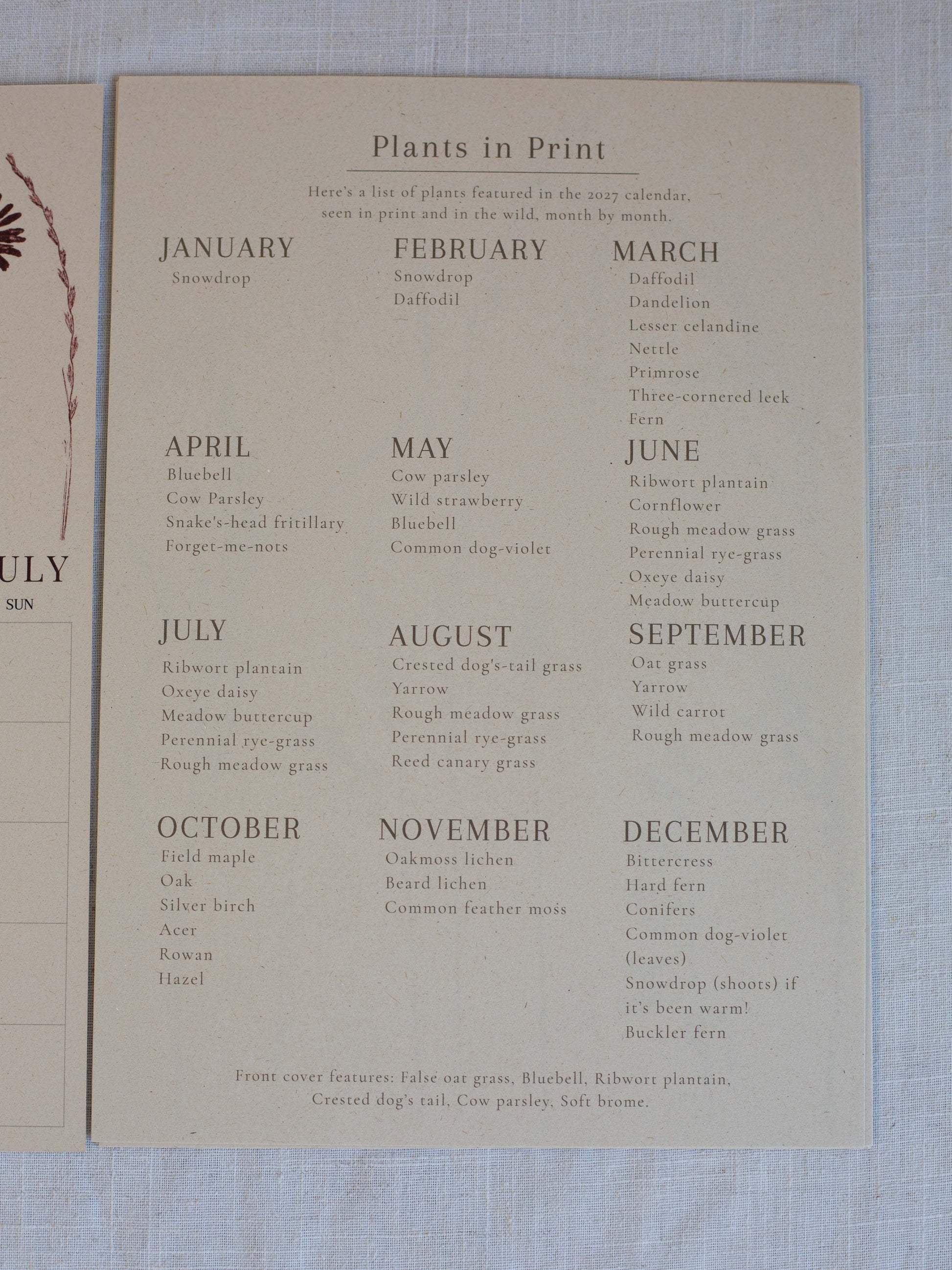 A vintage paper calendar page titled 'Plants in Print' laid on a light blue textured fabric background. The page lists months from January to December with plant names, and part of another paper sheet is visible on the left edge.