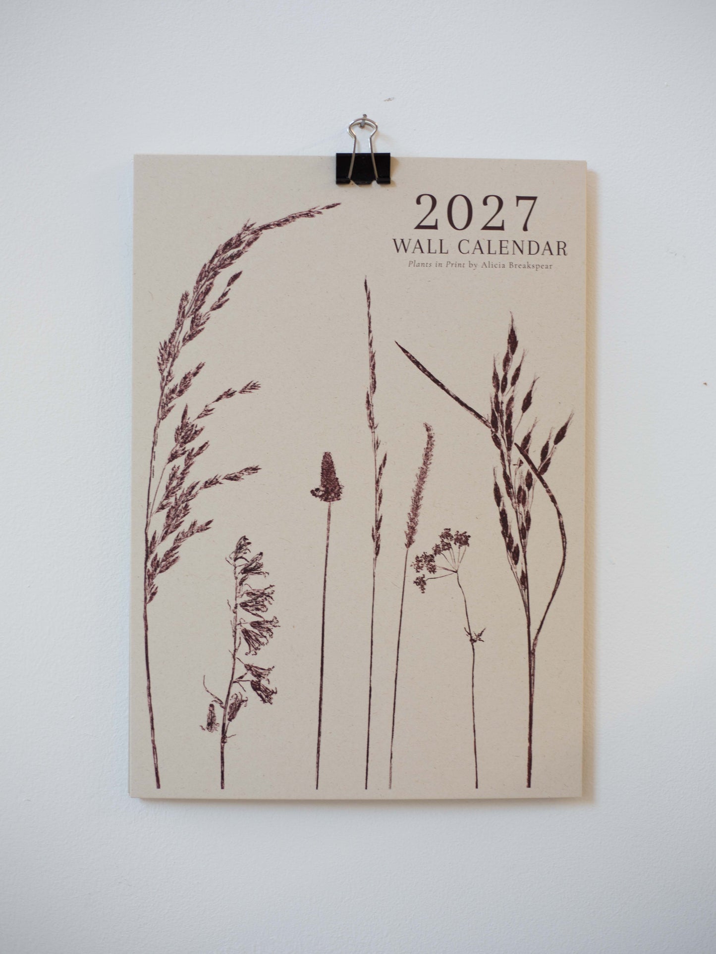 A 2027 wall calendar featuring botanical illustrations of wild grasses in dark brown on a warm beige background, hung on a white wall with a black binder clip.