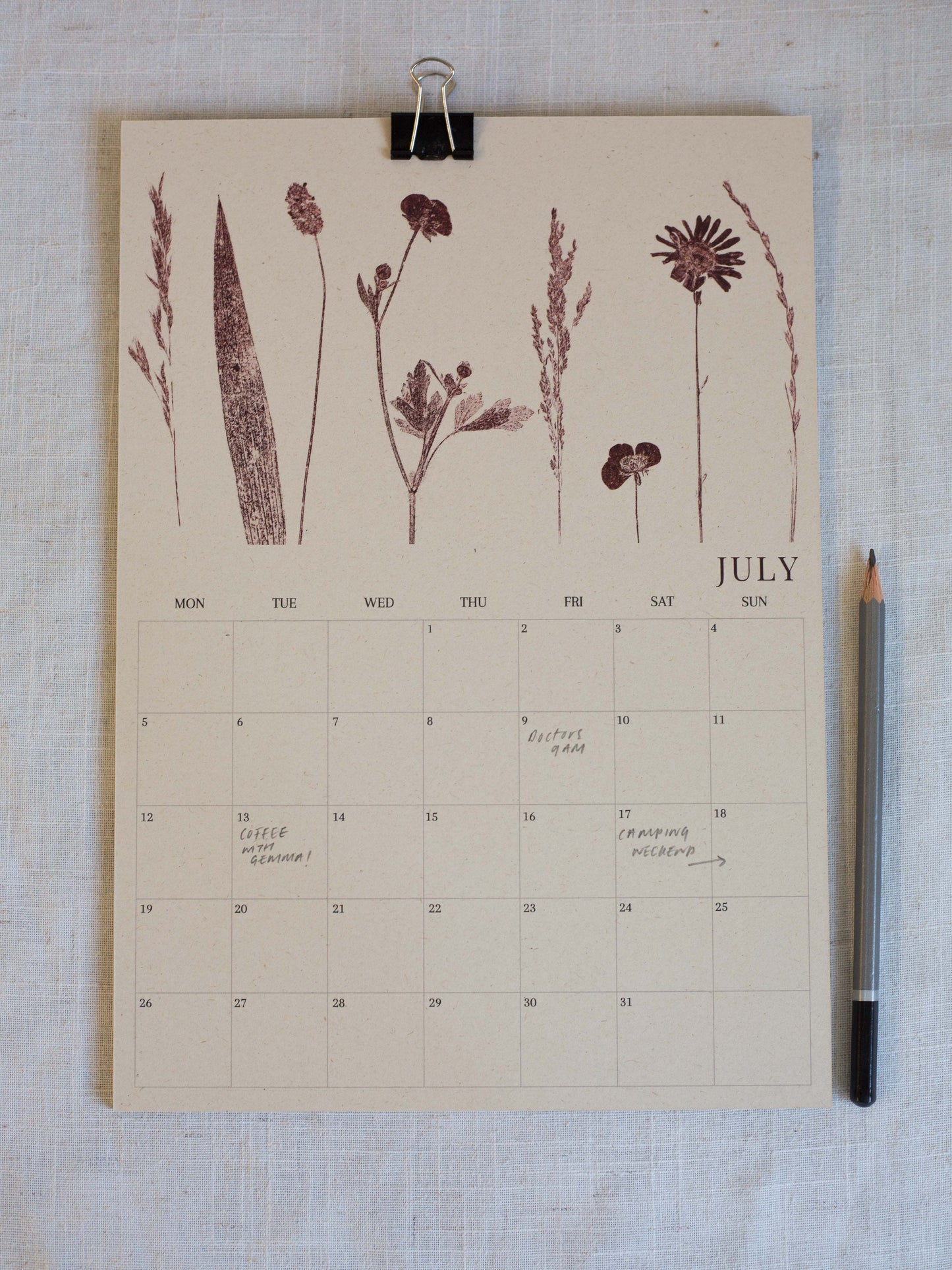 A July calendar page with botanical illustrations in dark brown at the top, photographed flat on a light fabric surface with a black binder clip at the top and a pencil on the right.