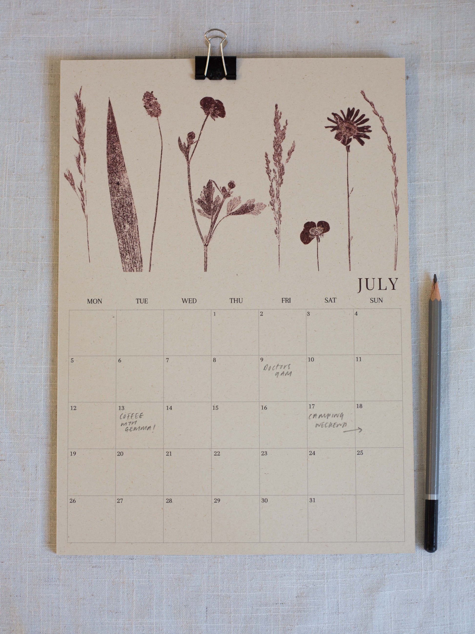A July calendar page with botanical illustrations in dark brown at the top, photographed flat on a light fabric surface with a black binder clip at the top and a pencil on the right.
